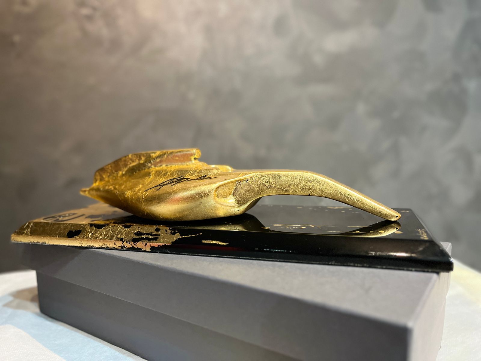 Escultura de mesa dourada da coleção AMK Emerson Fittipaldi, sobre base preta.